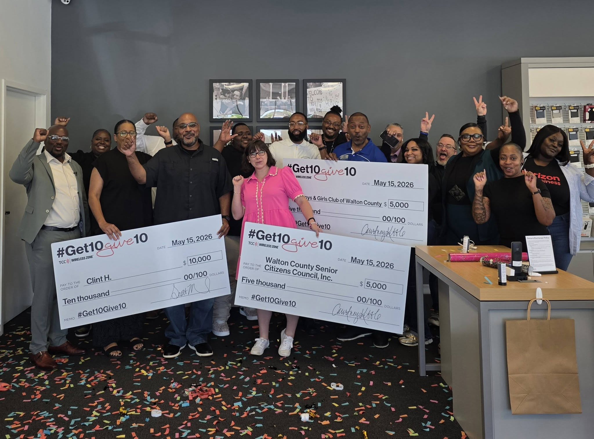 Group of people posing with oversized checks in an office, confetti on the floor from a celebration event.
